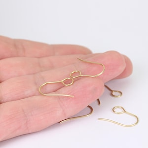 May include: Close-up of several gold-coloured earring hooks. The hooks have a simple, curved design with a small loop at the top and a hook at the bottom. They are likely used for making earrings. The hooks are displayed against a neutral background.