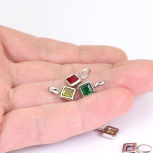 以下が含まれることがあります： カラフルな宝石が付いた、小さな正方形のペンダントのクローズアップ。各ペンダントには銀色のフレームとチェーン用のループが付いています。宝石は赤、緑、黄色です。ペンダントは明るい背景に表示されています。