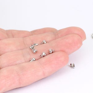 May include: A selection of small, silver crimp end beads, used in jewellery making. These metal findings have a rounded shape with a small loop for attaching to other components. The beads are scattered on a light background.