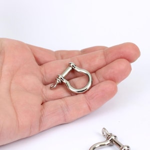 May include: A polished, silver shackle, a U-shaped metal fastener with a pin. The shackle is held in a hand against a white backdrop. This hardware is often used in marine and industrial applications.