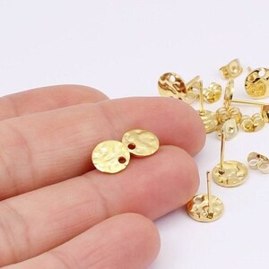 May include: Gold-colored earring components with a hammered texture. The image shows several round earring studs with posts and butterfly backs, along with two loose earring components. The components are small, with a textured surface and a hole for attachment.