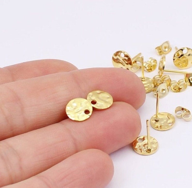 May include: Gold-colored earring components with a hammered texture. The image shows several round earring studs with posts and butterfly backs, along with two loose earring components. The components are small, with a textured surface and a hole for attachment.