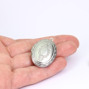 May include: Silver-colored oval locket with an ornate, engraved design. The locket has a decorative border and a small loop at the top for a chain. The locket is held in a hand against a white background.