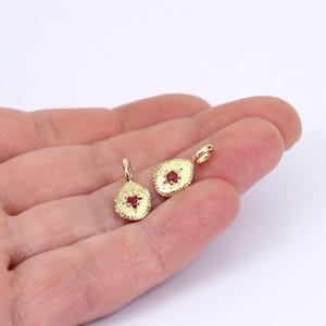 May include: Two gold-colored pendants with a textured oval design and a small red gemstone in the center. Each pendant has a small loop at the top for hanging. The pendants are displayed on a light background.