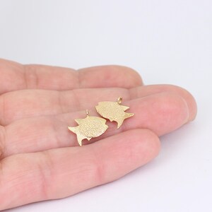 May include: Two small, gold-colored fish charms. Each charm has a textured surface and a small loop at the top for attaching to a chain or other jewelry component. The charms are displayed against a neutral background.