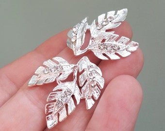 May include: Two silver leaf-shaped embellishments with clear rhinestones. The leaves are connected to each other and have a delicate, intricate design.