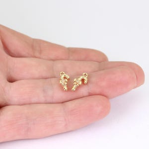 May include: Two small, gold-colored, abstract-shaped charms. Each charm has a curved, candy cane-like form with a cluster of small, round details. The charms are displayed against a neutral background.