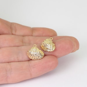 May include: Two small gold-toned metal charms shaped like fan-shaped seashells. Each charm features a textured, ribbed surface and a small circular loop at the top for attachment. The metallic finish is reflective and bright, highlighting the shell details.