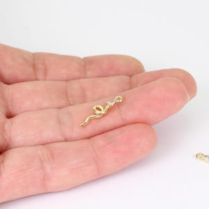 May include: A gold-coloured charm in the shape of a stylised ribbon with a small, clear gemstone at the top. The charm has a loop for attaching to jewellery and is shown against a light background.