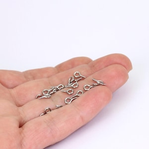 May include: A selection of small, silver-coloured screw eye pins. Each pin has a looped eye and a threaded screw end. The pins are spread across a light background, indicating their use in jewellery making or crafts.