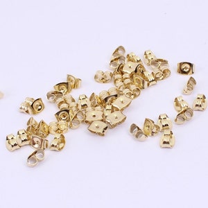 May include: A group of gold-coloured earring backs scattered on a white surface. The backs are designed to secure earrings to the earlobe.