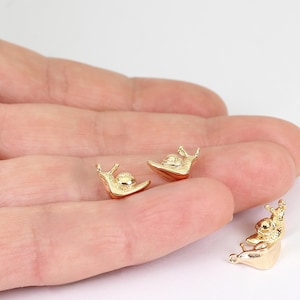 May include: Three small, gold-toned metal snail charms with detailed shell textures and smooth, curved bodies. Each snail charm features a small loop at the top of the shell, designed for attachment to jewellery or other decorative craft projects.