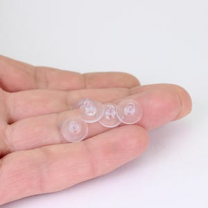 May include: A close-up of several clear, round earring backs. The earring backs are made of a translucent material and have a flat, disc-shaped base with a small post in the center. They are designed to secure earrings.