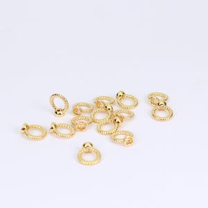 Könnte beinhalten: Eine Sammlung kleiner, goldfarbener Schmuckzubehörteile. Jedes Zubehörteil hat eine Ringform mit einem strukturierten Muster und einer kleinen, runden Perle oben. Die Zubehörteile sind auf einer weißen Oberfläche verstreut.
