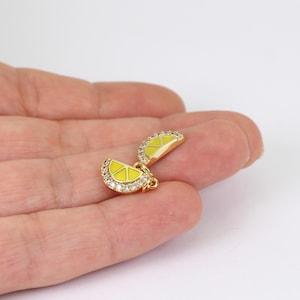 May include: A gold-toned charm shaped like a lemon slice, featuring bright yellow enamel detailing and a border of small, clear, sparkling stones. The charm has a small metal loop at the top for attachment, showcasing a vibrant citrus design.