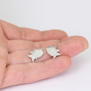 May include: Two silver-colored fish-shaped charms. Each charm has a textured surface and a small loop at the top for attaching to a chain or bracelet. The charms are displayed against a light background.