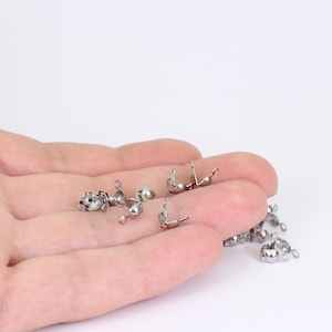 May include: A collection of small, shiny, silver-colored jewelry findings, including crimp beads and end caps. These components are used in jewelry making to secure beads and attach clasps. The findings are displayed against a white background.