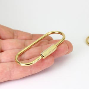 May include: A gold-colored, oval-shaped carabiner with a hexagonal screw-lock mechanism. The carabiner is held in a hand against a white background. The metal has a polished, reflective surface.