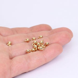 May include: A collection of small, shiny gold-colored ball-end findings, used for jewelry making. The findings are round with a loop for attaching to other components. The findings are held in a hand against a white background.