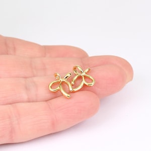 May include: Two small, gold-colored bow-shaped charms. The charms are made of a shiny metal and have a delicate, open design. They are likely jewelry components or decorative elements for crafts.