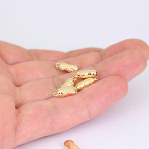 以下が含まれることがあります： 小さな金色のピーナッツチャームがいくつか表示されています。チャームには溝が詳細に施されており、片方の端に小さなループが付いています。ピーナッツは白い背景に対して手で持たれています。これらのチャームは、ジュエリー作りなどに使用できます。