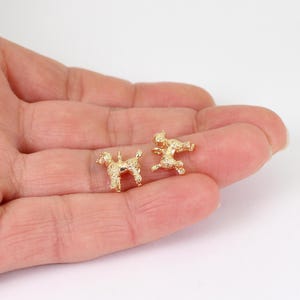 May include: Two small, gold-coloured poodle charms. One charm is standing, the other is in a playful pose. The charms are detailed with textured fur and are likely made of metal. They are displayed on a light background.