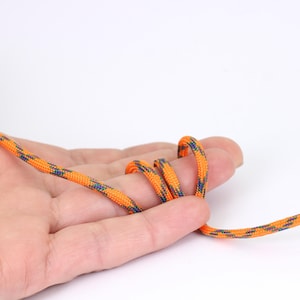 May include: Close-up of a length of orange and blue patterned rope. The rope is looped over a person's fingers. The rope has a repeating pattern of orange, blue, and green. The background is white.