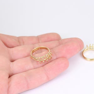 May include: Gold-colored adjustable ring with a decorative design featuring multiple small loops. The ring is shown on a hand against a white background. Another similar ring is visible in the background.