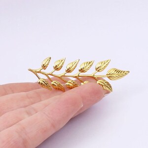 May include: A gold-toned leaf branch brooch with a delicate design. The brooch features multiple leaves arranged in a flowing pattern.