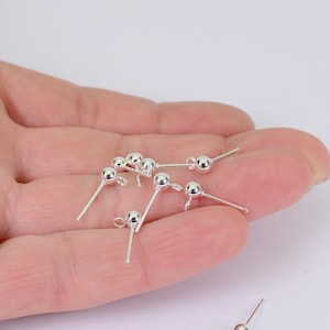 May include: A collection of small, silver-colored earring posts with a ball-shaped design. Each post has a small loop and a straight pin for insertion. The earrings are displayed on a light background.