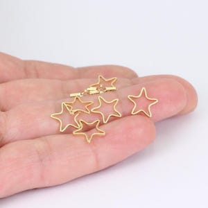 May include: Gold-colored star-shaped charms. The charms are open-frame, with a five-pointed star design. Several charms are visible, some scattered and others connected with a clasp. The background is a plain white.