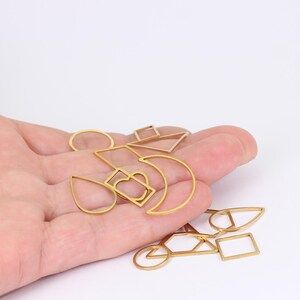 May include: A selection of gold-coloured metal charms in diverse geometric forms, such as a circle, a square, a teardrop, a crescent moon, and a bicycle. The charms are held in a hand against a white backdrop. These charms are suitable for jewellery making.