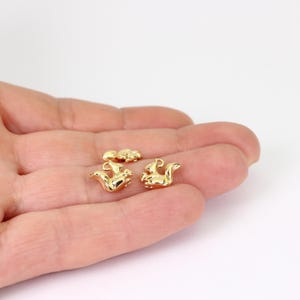 May include: Small, gold-coloured squirrel charms, likely for jewellery making, are shown in a person's hand. The charms feature a detailed squirrel design. The background is plain white, which highlights the charms.