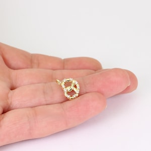 May include: A gold-colored pretzel charm with small, clear gemstones. The charm is in the shape of a pretzel, with the gemstones set around the loops. The charm is small and delicate, and is likely a pendant or charm for a necklace or bracelet.