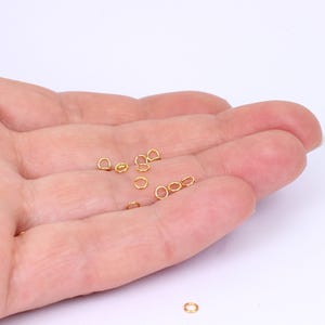 May include: A close-up of a hand holding a pile of small, gold jump rings. The rings are approximately 5mm in diameter and are ideal for jewellery making.
