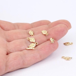 Könnte beinhalten: Eine Sammlung kleiner, goldfarbener Metallanhänger in Blattform, die jeweils ein detailliertes Adern-Muster und eine kleine kreisförmige Öse an der Oberseite zur Befestigung aufweisen. Diese metallischen Blatt-Elemente liegen verstreut.