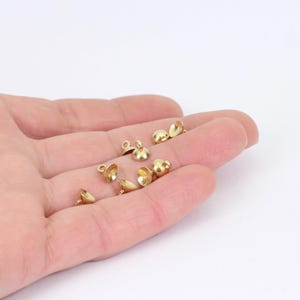 May include: A close-up of a hand holding several gold-coloured metal bead caps. The caps are small and round, with a loop on top for attaching to a string or chain.