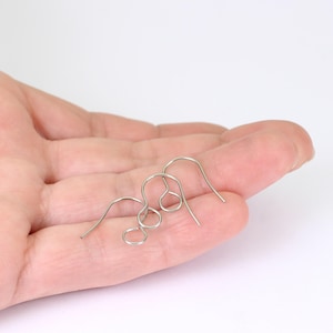 May include: Three silver earring hooks are displayed. The hooks are made of thin wire and have a curved design. The hooks are held in a person's open hand against a white background.