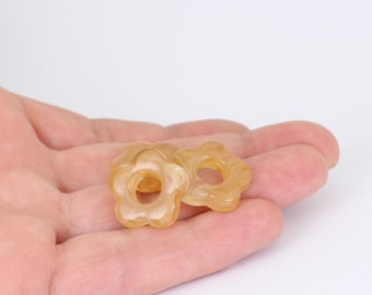 May include: Three translucent, flower-shaped beads in shades of yellow and cream are displayed. The beads have a hole in the center and are held in a person's hand against a white background. The beads have a glossy finish.