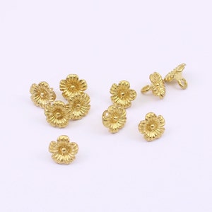May include: A selection of small, gold-coloured flower-shaped beads. Each bead features detailed petal and centre designs, ideal for jewellery making or craft projects. The beads are scattered on a white surface.