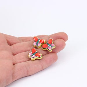 May include: Three small, colorful butterfly figurines. Each butterfly has a yellow base, red and pink wings, and a green accent. The figurines are held in a person's hand against a white background.