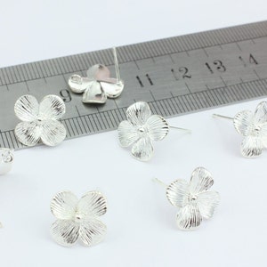 May include: Silver flower stud earrings with detailed petal design. The earrings have posts for pierced ears. Several earrings are shown, with a ruler in the background for scale.
