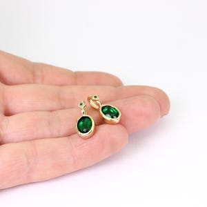 May include: Two gold-colored pendant earrings with oval, emerald-green gemstones. Each earring has a small, square-shaped accent stone above the oval gem. The earrings are displayed on a light background.