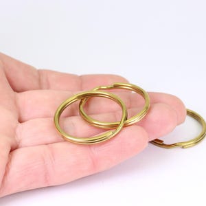 May include: Three brass key rings, two interlocked, are held in a hand against a white background. The key rings are a warm gold colour, and the metal has a smooth, reflective surface. The rings are designed to hold keys or other small items.