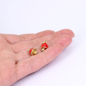 以下が含まれることがあります： 赤い帽子と白い水玉模様の、小さな金色のキノコのチャームが2つ。白い背景の中で、人の手の中にチャームが収まっている。キノコの茎は明るい金色です。