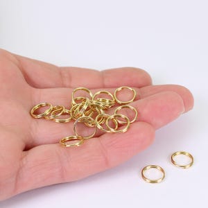 May include: A selection of small, gold-coloured split rings, displayed on a light surface. These double-loop rings are suitable for jewellery making or crafts. The rings are made of a shiny, metallic material.