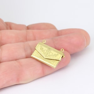 May include: A small, gold-colored envelope-shaped charm with intricate floral and leaf patterns. The charm has two small loops at the top for attaching to a necklace or bracelet. The charm is held in a hand.