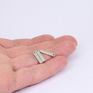 May include: Three small, rectangular, silver-colored metal charms with a textured surface. Each charm has a small hole at one end. The charms are held in a hand against a white background.