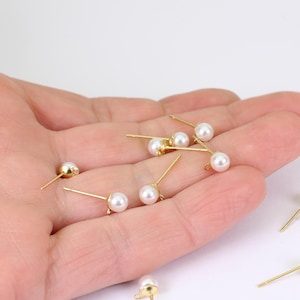 May include: A selection of small, gold-coloured stud earrings, each featuring a white, round pearl. The earrings are presented against a light backdrop, highlighting their delicate design. The earrings are small and elegant, suitable for everyday wear.
