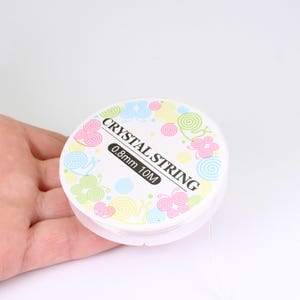 May include: A spool of clear crystal string, 0.8mm thick and 10 meters long, is displayed. The white spool has a decorative label with the words "Crystal String" and colorful spiral designs in pink, yellow, green, and blue.